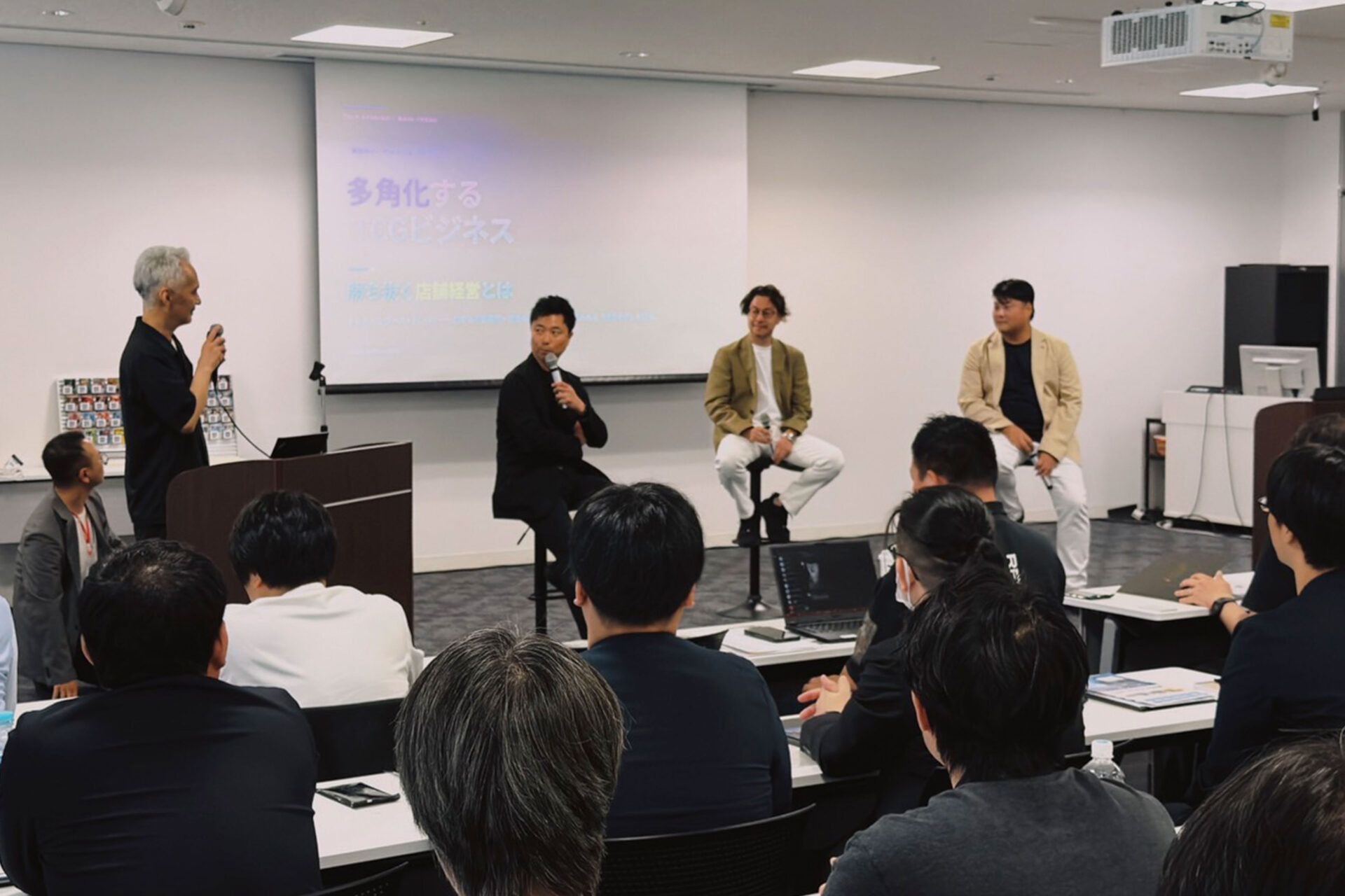 【アフターレポート】満員御礼!「第2回 次世代TCGショップカンファレンス」 2 「多角化するTCGビジネス」というテーマが映し出されたスクリーンの前で、マイクを持って話す登壇者と、ステージ上で耳を傾ける3名のスピーカー、手前でセッションを聴講する参加者たちの様子。